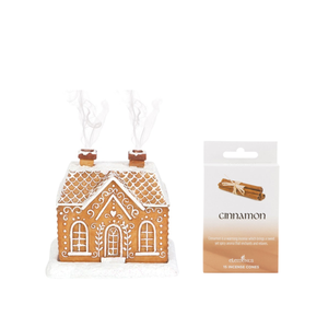 A gingerbread house-shaped incense holder with white icing details and two small brown incense sticks sticking out of the roof, shown next to its retail packaging card labeled 'cinnamon' by elvetkin.