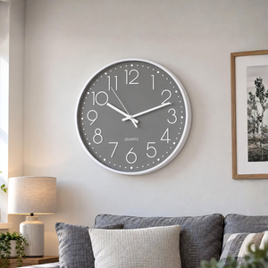 A round gray quartz wall clock with a white frame and white numerals, mounted on a light beige wall above a gray sofa in a modern living room setting.