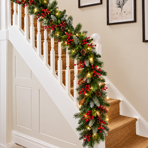 A festive Christmas stair garland with green pine branches, red berries, white faux snow, and warm white string lights draped over a white staircase railing in a home interior.