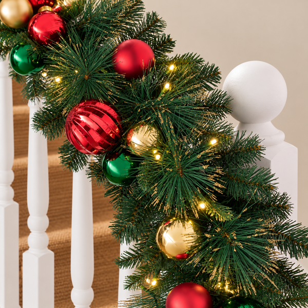 A close-up of a green Christmas garland with red, green, and gold ornaments and warm white string lights draped over a white wooden staircase railing.