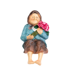 A ceramic garden figurine of a smiling woman sitting barefoot and holding a bouquet of pink flowers, set against a plain white background.