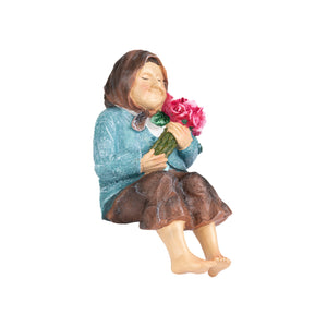 A small figurine of a seated child holding a bouquet of pink flowers, wearing a textured blue top and brown skirt, isolated on a white background.