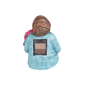A garden statue of a woman with brown hair, wearing a textured light blue dress, shown from the back. A rectangular solar panel is mounted on the back of the statue.