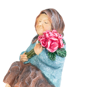 A decorative figurine of an elderly woman with closed eyes, wearing a brown headscarf and a textured light blue dress, holding a large bouquet of pink roses with green leaves, isolated on a white background.