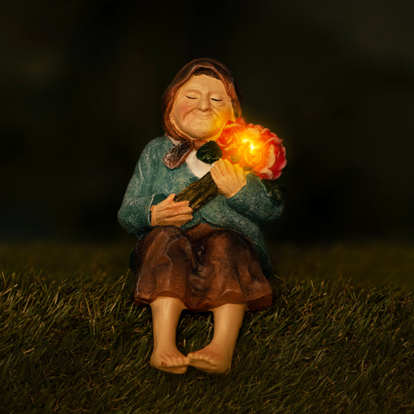 A small seated figurine of a woman holding a glowing flower, photographed outdoors on grass at night. The figurine has a blue-green top, brown skirt, and bare feet, with the flower emitting a warm orange light.