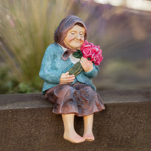 A small decorative figurine of an elderly woman sitting barefoot on a stone wall, holding a bouquet of pink flowers. The figurine has a weathered, rustic finish with a teal-blue top, brown skirt, and purple headscarf, set against a blurred outdoor background.