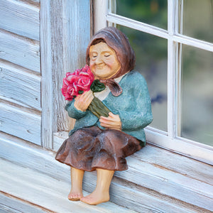 A decorative figurine of an elderly woman sitting on a weathered wooden windowsill, holding a bouquet of pink roses. The figurine has a textured teal-blue jacket, a brown skirt, and a brown head covering, set against a rustic blue-gray wooden wall and window frame.