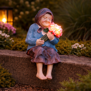 A decorative garden figurine of an elderly woman sitting on a stone wall, holding a glowing lighted rose bouquet. The figurine is dressed in a blue top and a brown skirt, set against a blurred garden background with plants and a warm lantern.