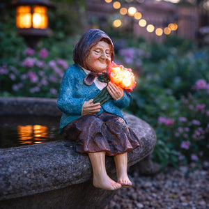 A decorative garden statue of a smiling woman in a blue dress and brown skirt sitting on a stone edge, holding a glowing light bulb shaped like a rose, with a blurred garden background and warm string lights.