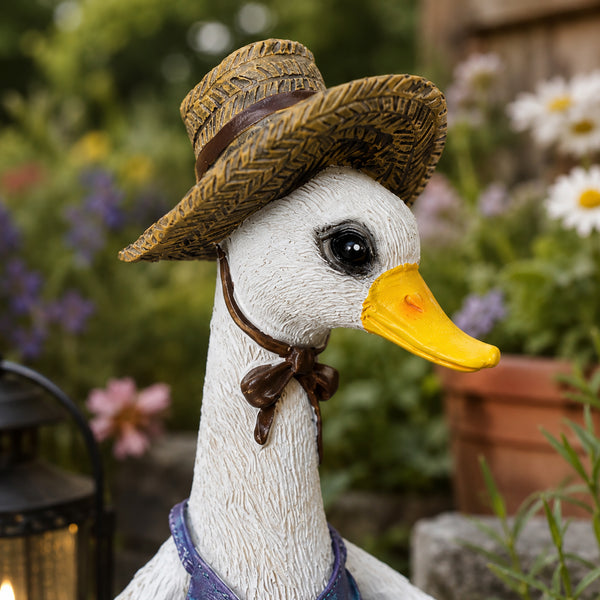 A decorative garden duck figurine with a white neck and yellow beak, wearing a woven straw hat with a brown band and bow tie, set against a blurred garden background.