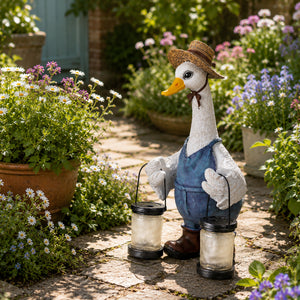 A decorative garden statue of a white goose wearing a straw hat, blue overalls, and brown boots, holding two black lanterns on a stone pathway surrounded by potted flowers and greenery.