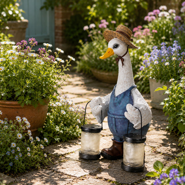A decorative garden statue of a white goose wearing a straw hat, blue overalls, and brown boots, holding two black lanterns on a stone pathway surrounded by potted flowers and greenery.