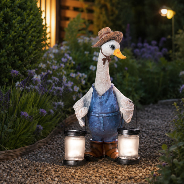 A decorative garden statue of a goose dressed as a farmer, wearing a brown hat, blue overalls, and brown boots, holding two lit lanterns on a gravel path at night with garden plants in the background.