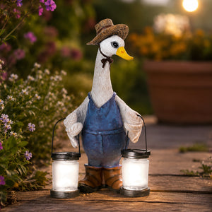 A decorative garden duck figurine wearing a brown hat and blue overalls, holding two glowing lanterns on a wooden path with a blurred garden background.
