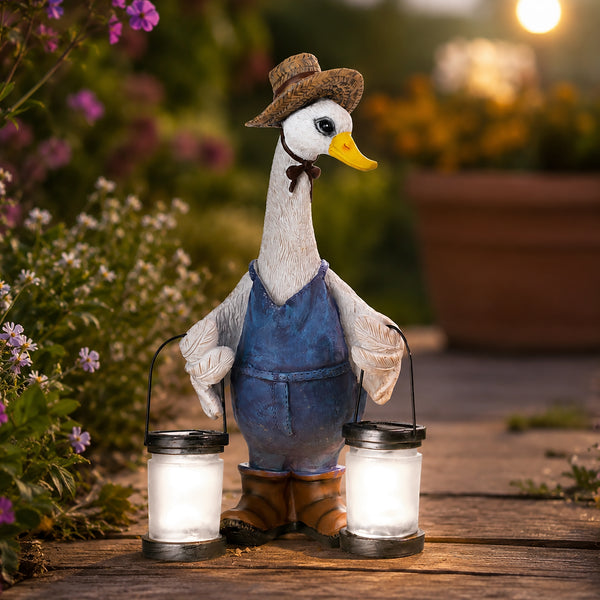 A decorative garden duck figurine wearing a brown hat and blue overalls, holding two glowing lanterns on a wooden path with a blurred garden background.