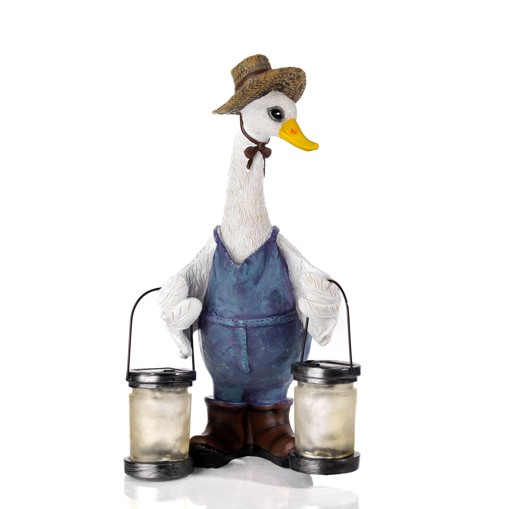 A decorative garden statue of a white goose wearing a straw hat, blue overalls, and brown boots, holding two glowing lanterns on a stone path with plants in the background.