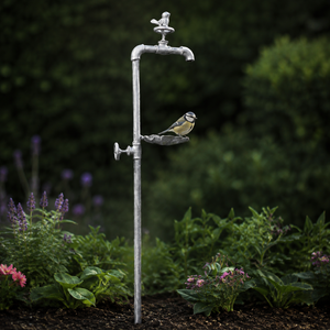 A decorative garden stake shaped like an old silver faucet with a small bird perched on the spout, set in a garden with green foliage and flowers.