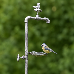 A small bluebird perched on a metal garden bird feeder shaped like an old faucet, with a glass bowl attached below the spout, set against a soft blurred green garden background.