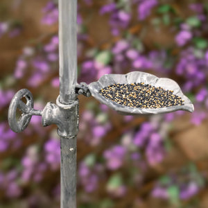 A leaf-shaped metal bird feeder filled with mixed birdseed, attached to a garden fence post in an outdoor setting with a blurred background of purple flowers.