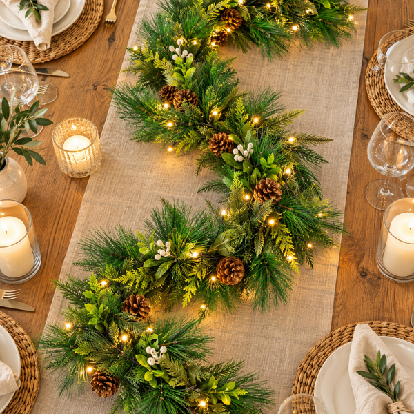 A festive Christmas table runner garland made of green pine branches, brown pinecones, and warm white string lights, laid on a beige burlap table runner over a wooden dining table with place settings and candles.