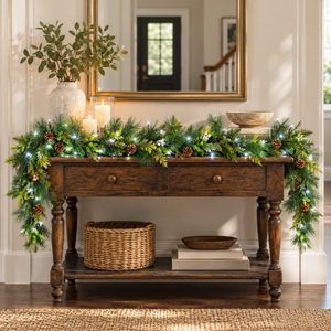 A dark wood console table decorated with a green pine garland lit with white fairy lights, accented with pinecones, in a warm, festive interior setting.