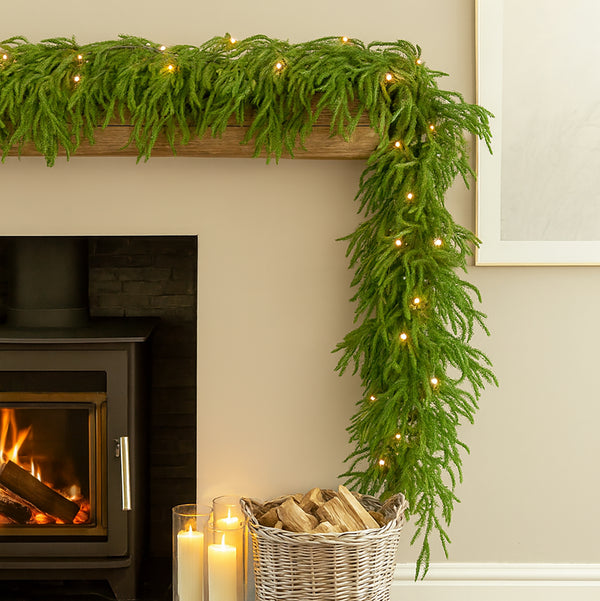 Decorative green garland with lights on a fireplace mantel, candles, and firewood basket.