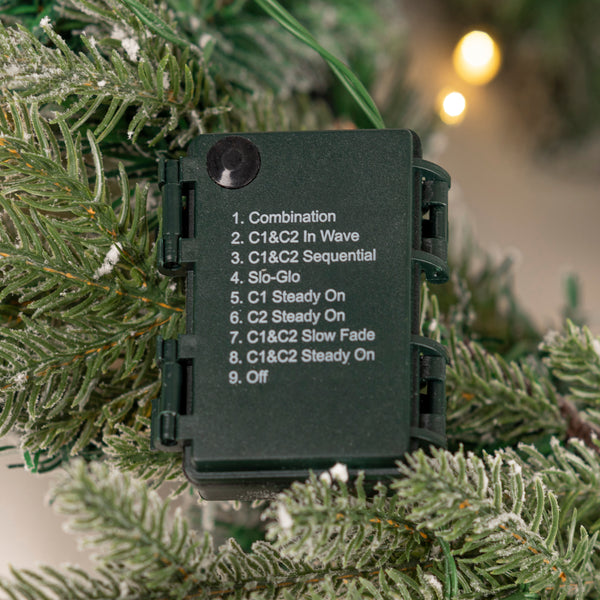 A black rectangular electronic device labeled with nine settings including Combination, C1&C2 In Wave, C1&C2 Sequential, Si&Glo, C1 Steady On, C2 Steady On, Slow Fade, and Off, shown hanging on a frosted Christmas tree branch with warm bokeh lights in the background.