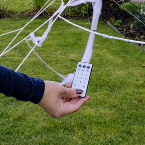 A hand holding a white remote control with a black frame, positioned in front of a white cobweb garden net set up on a green lawn.