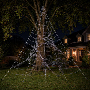 A large white LED spiderweb light fixture shaped like a pyramid is strung on a tree in a backyard at night, with a house and lawn visible in the background.