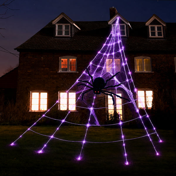 A large decorative spider web Halloween decoration with glowing purple LED webbing and a black spider in the center, displayed outdoors in front of a house at night.