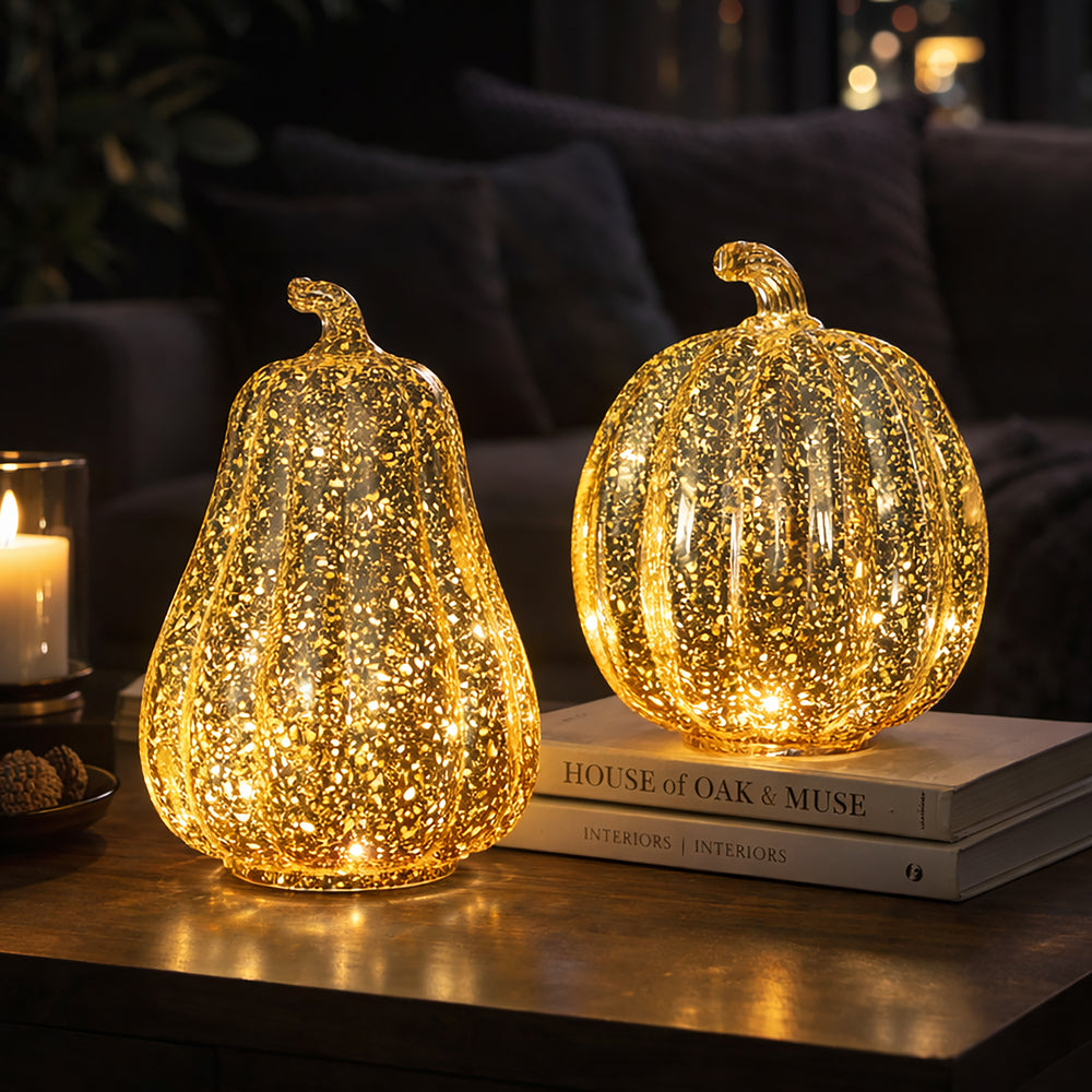 Two decorative glass pumpkins with warm white LED lights inside, one pear-shaped and one round, displayed on a wooden table in a cozy living room setting.