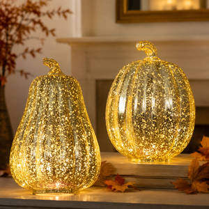 Two speckled glass effect pumpkins with warm yellow internal lighting, one pear-shaped and one round, displayed on stacked books with autumn leaves on a mantel.