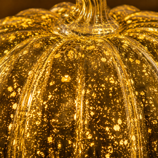 A close-up of a decorative glass pumpkin with a ribbed shape, glowing with warm golden light and filled with tiny reflective sparkles.