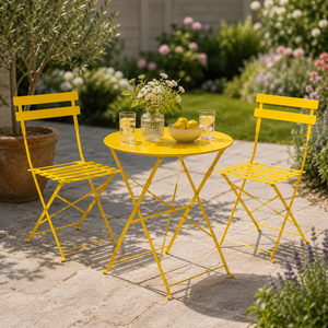A bright yellow outdoor bistro set consisting of a round folding table and two matching folding chairs, arranged on a stone patio in a garden setting with a vase of flowers and glasses of lemonade.