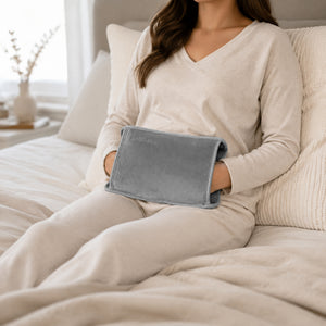 A woman in a cream-colored pajama set sits on a bed holding a gray heated pad against her lower back. The room has a soft, neutral color palette with beige and white tones.