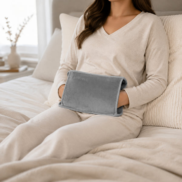 A woman in a cream-colored pajama set sits on a bed holding a gray heated pad against her lower back. The room has a soft, neutral color palette with beige and white tones.