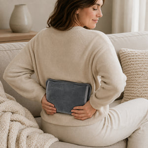 A woman sitting on a beige couch, viewed from behind, holding a dark gray heated pad against her lower back. She is wearing a cream ribbed knit sweater and matching pants in a cozy living room setting.