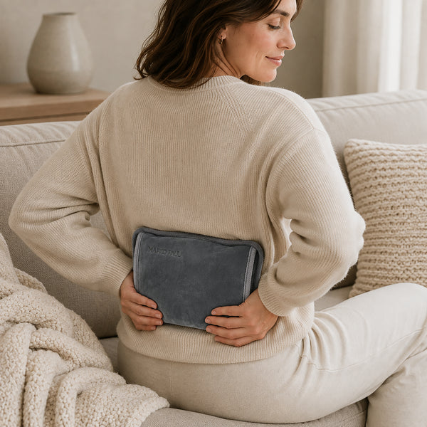 A woman sitting on a beige couch, viewed from behind, holding a dark gray heated pad against her lower back. She is wearing a cream ribbed knit sweater and matching pants in a cozy living room setting.