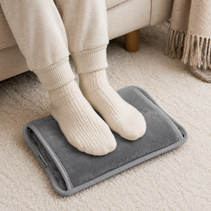 A gray heated foot rest pad on a beige carpet with a person's feet in cream knit socks resting on it, near a sofa and throw blanket.