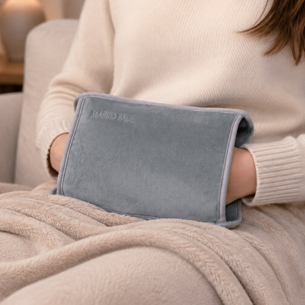 A person in a cream knit sweater holds a light blue-gray plush hand warmer with the brand name 'MARIO BAIL' embroidered on it, wrapped in a beige blanket on a couch.