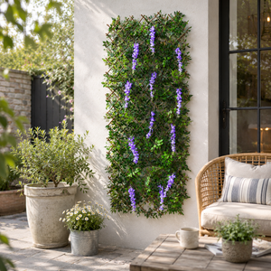 A vertical green wall garden with purple wisteria flowers hanging from a wooden trellis, styled on a white stucco wall in a patio setting with a wicker chair, potted plants, and a stone table.