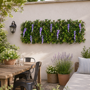 A rectangular green artificial hedge wall with hanging purple wisteria-style flowers mounted on a beige stucco wall in a garden setting, with a wooden table, chairs, potted plants, and a cushion nearby.
