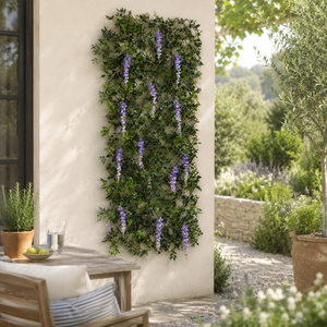 A vertical artificial wisteria hedge with cascading purple flowers mounted on a light beige stucco wall in a sunny outdoor patio setting.