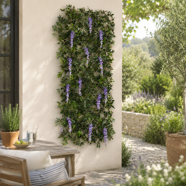 A vertical artificial wisteria hedge with cascading purple flowers mounted on a light beige stucco wall in a sunny outdoor patio setting.