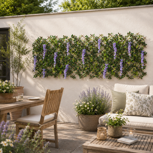 A styled outdoor patio scene with a white wall decorated with a green trellis covered in purple wisteria flowers. In the foreground, a wooden dining chair, dining table, coffee table, and a cream sofa with patterned pillows are arranged in a garden setting.
