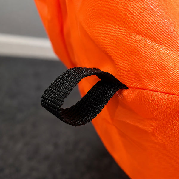 Close-up of a bright orange fabric item with a black webbing loop handle, photographed indoors against a dark gray floor and light gray wall.