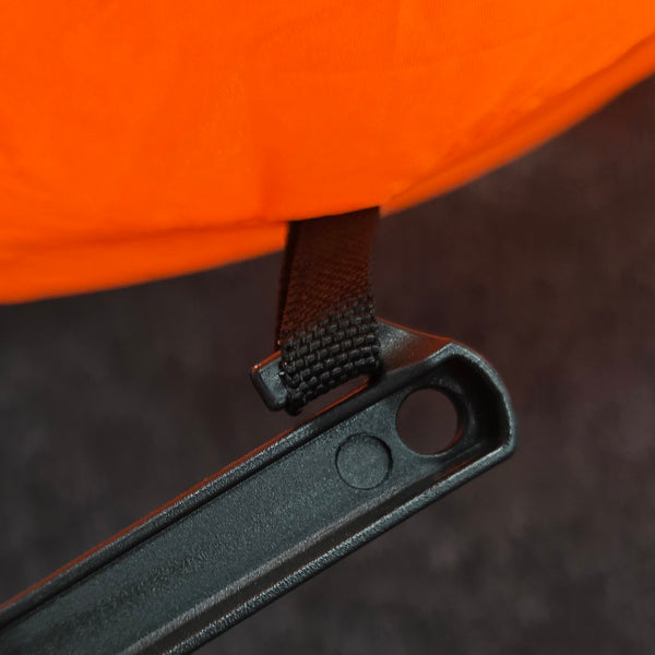 Close-up detail of an orange tent fabric attached to a black plastic hanging loop with a dark brown webbing strap, set against a dark textured background.