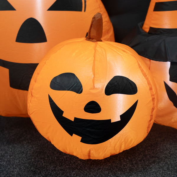 An inflatable orange jack-o'-lantern pumpkin with a black face and stem, photographed outdoors on a dark ground with other similar pumpkins partially visible behind it.
