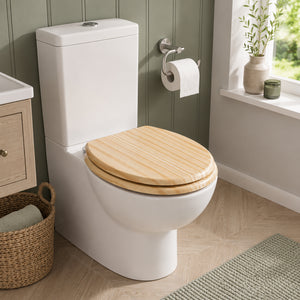 A white ceramic toilet with a light natural wood seat lid, shown in a styled bathroom setting with green paneled walls, a window, and a woven basket.