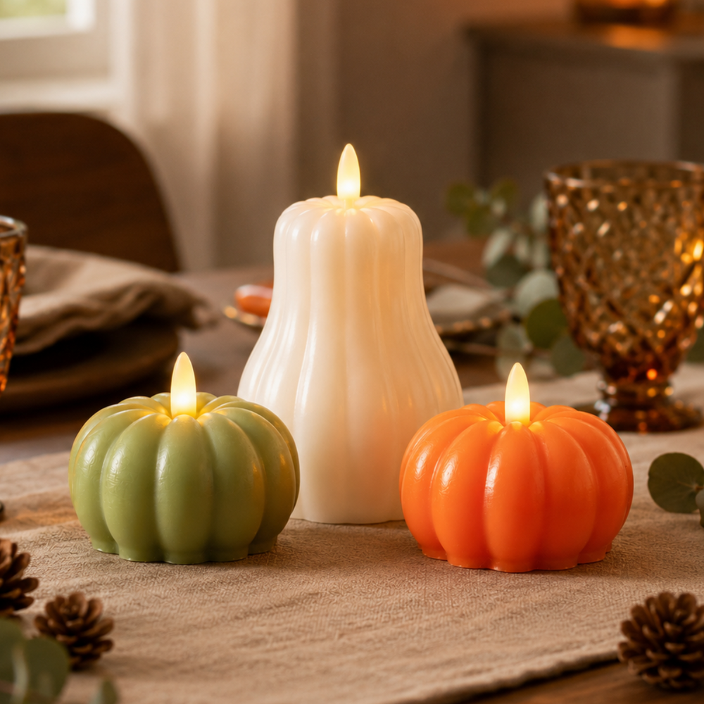 Three flameless LED pumpkin-shaped candles in green, white, and orange are arranged on a burlap table runner with eucalyptus leaves and pinecones in a warm, cozy dining setting.